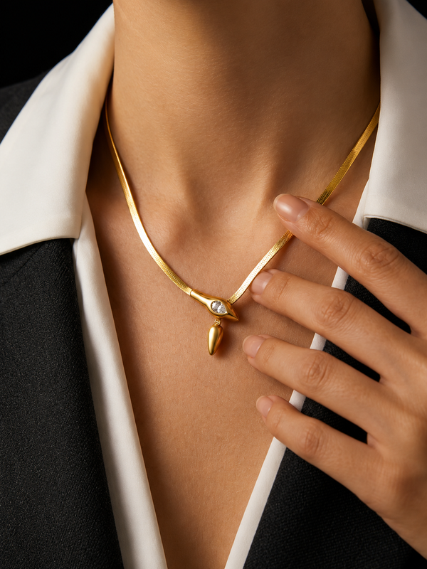 A close-up of a model wearing a gold snake-necklace with a small clear stone and teardrop pendant, styled with a black and white collared shirt.