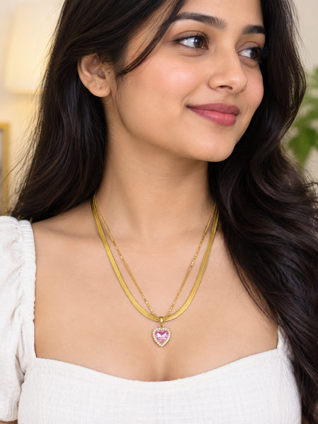 A woman wearing a layered gold necklace with a heart-shaped pink and white stone pendant, shown in a close-up portrait.