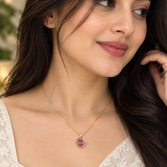 A woman wearing a delicate gold chain necklace with a heart-shaped pendant featuring a pink gemstone surrounded by small clear stones, shown in a close-up portrait.