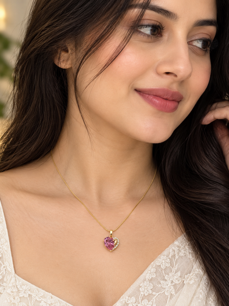 A woman wearing a delicate gold chain necklace with a heart-shaped pendant featuring a pink gemstone surrounded by small clear stones, shown in a close-up portrait.