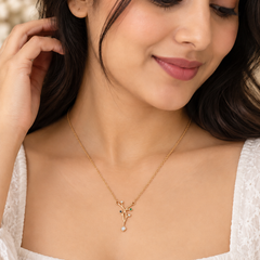 A woman wearing a delicate gold necklace with a small leafy branch pendant and a tiny light blue bead, paired with a white lace top.