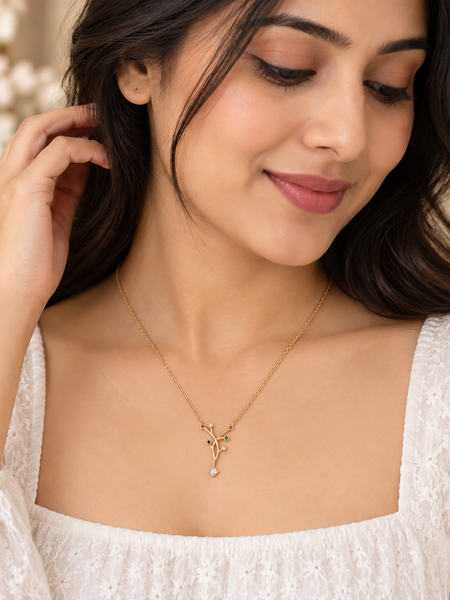 A woman wearing a delicate gold necklace with a small leafy branch pendant and a tiny light blue bead, paired with a white lace top.